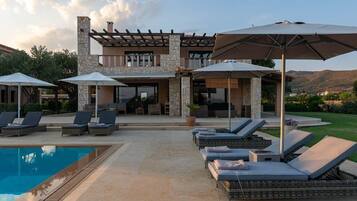 A poolside area with lounge chairs and umbrellas, a stone building with a pergola, and a view of the mountains.