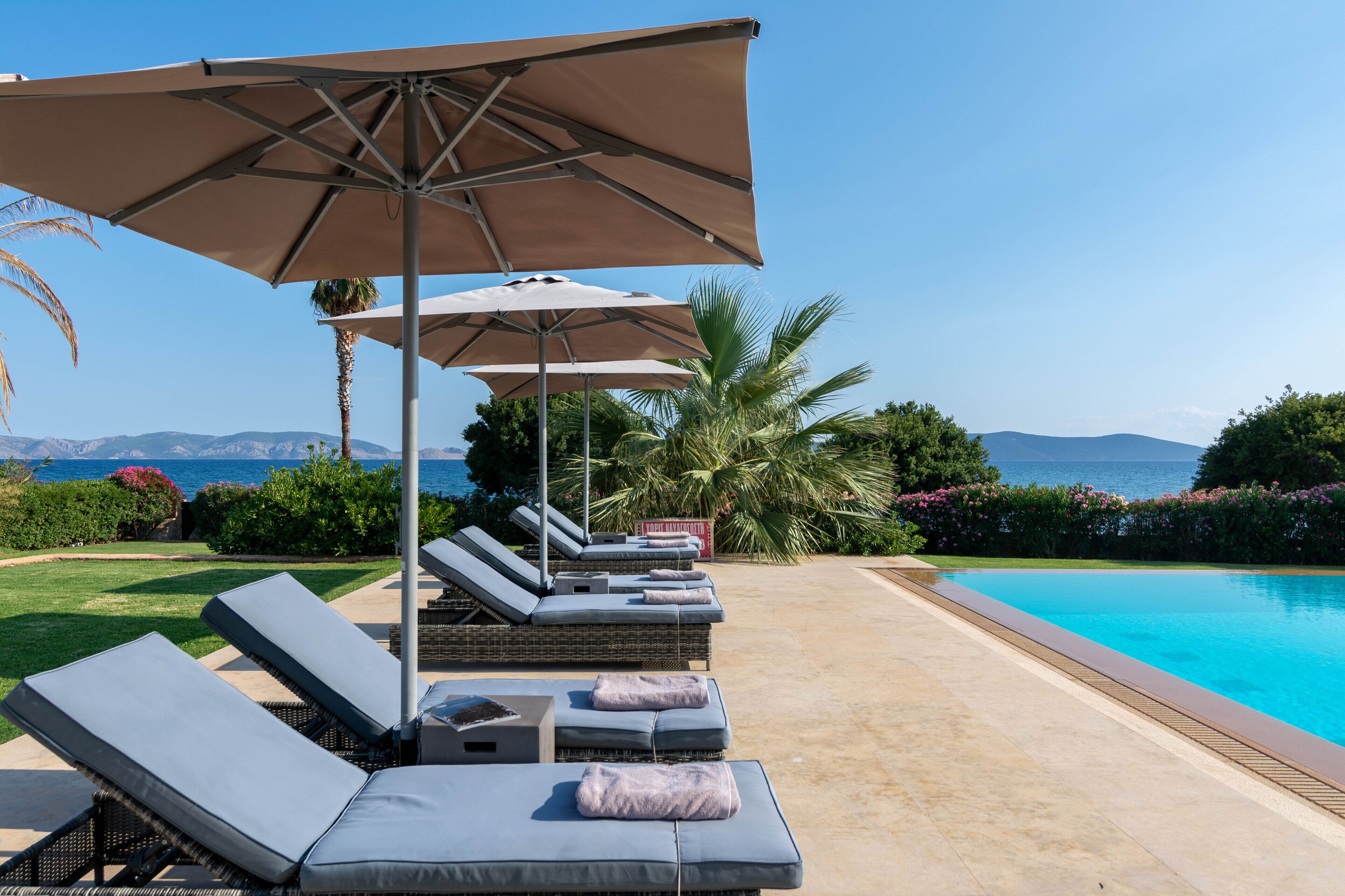 Ein Poolbereich mit Liegestühlen und Sonnenschirmen, mit Blick auf das klare Meer und einem gepflegten Garten.