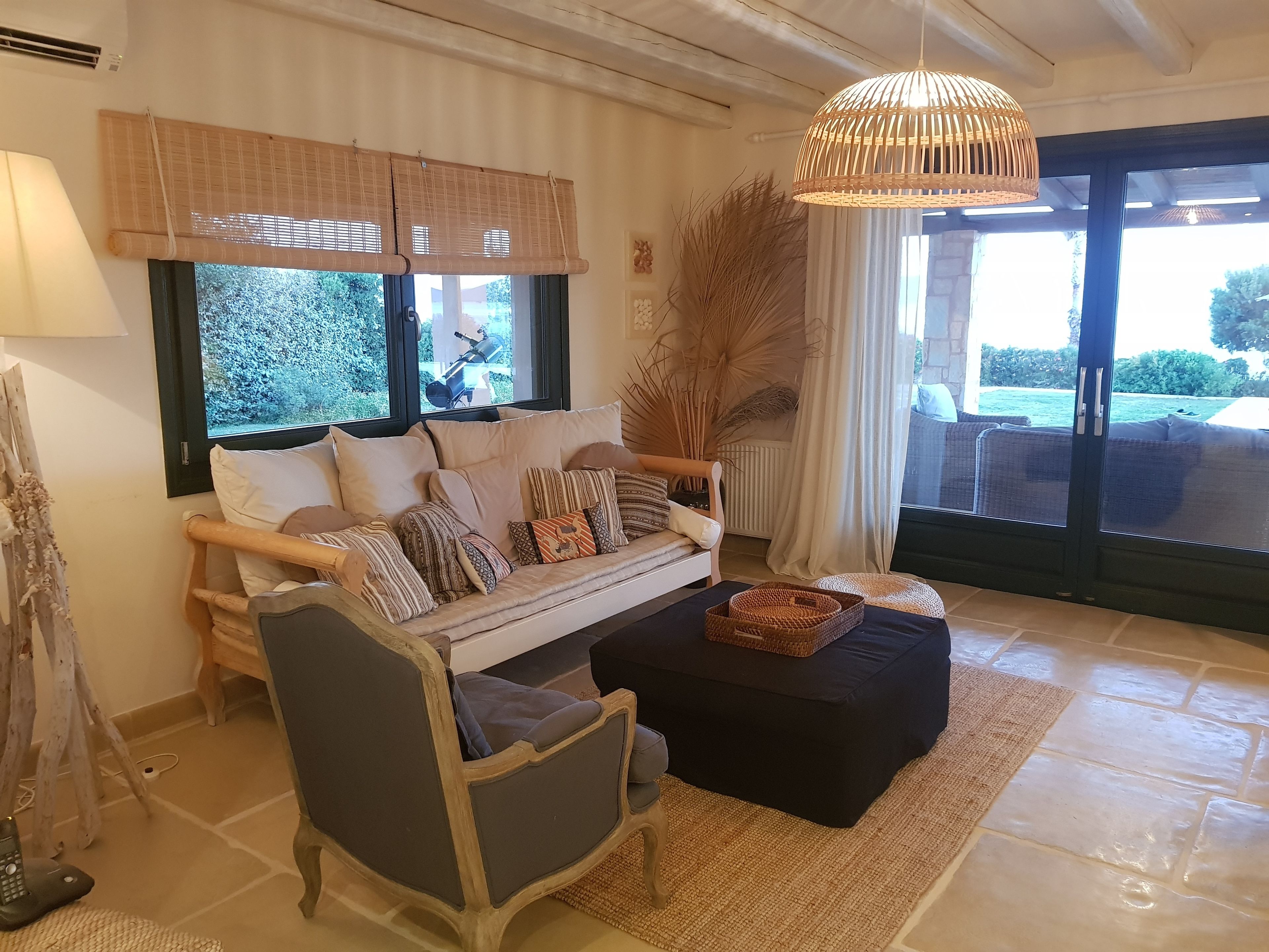A cozy living room with a sofa, armchair, and coffee table, large windows with blinds, and a hanging light fixture.