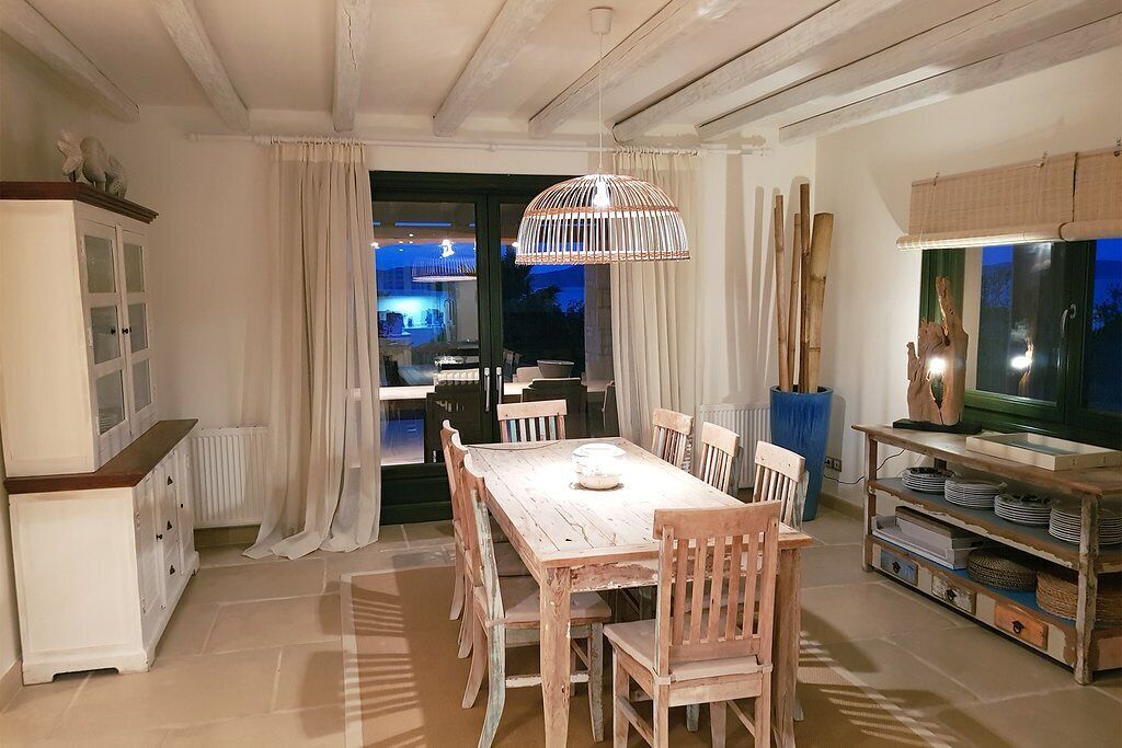A dining area with a wooden table and chairs, a hanging light fixture, and a view into another room.