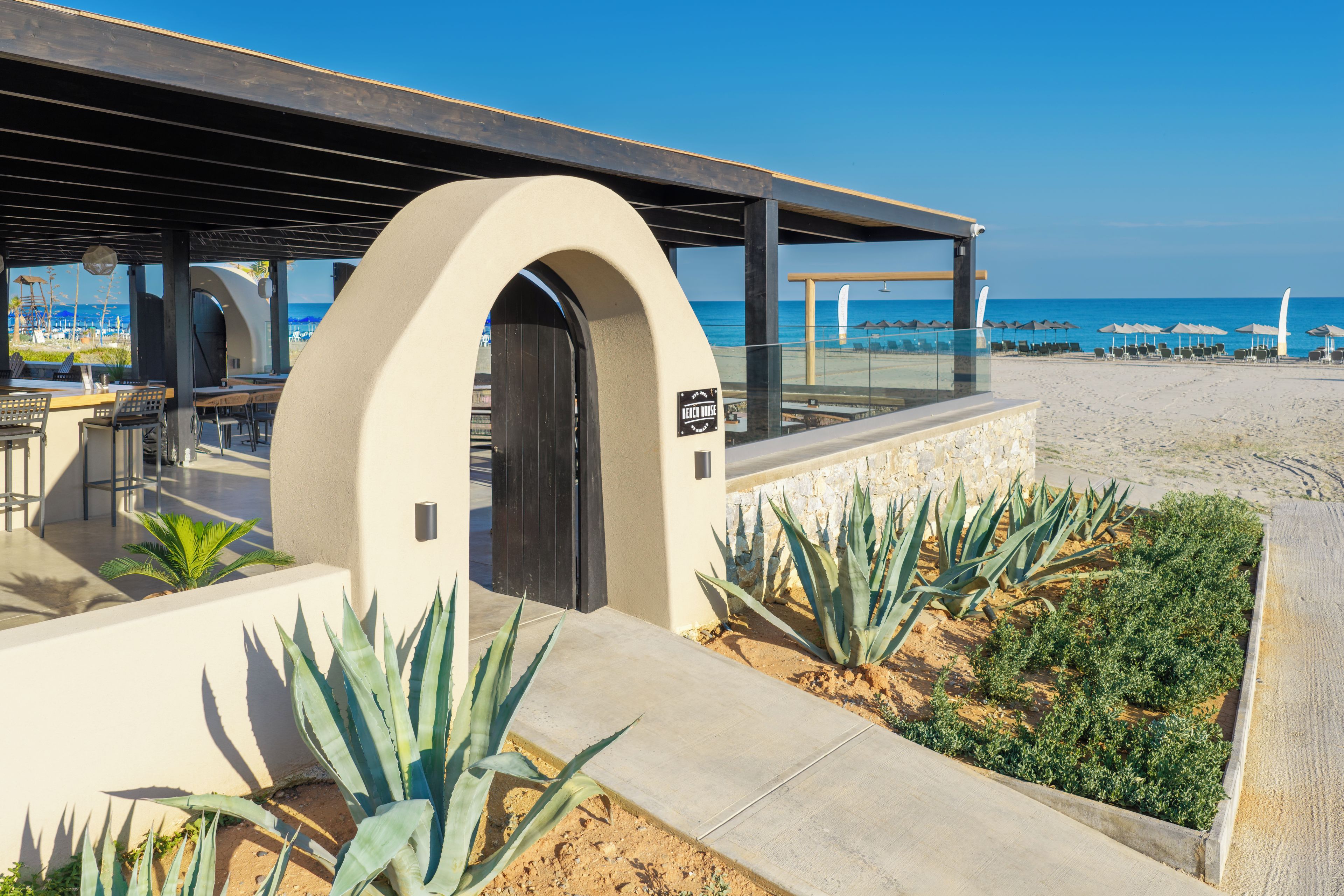 A beachside restaurant with outdoor seating, a view of the ocean, and a sign indicating 'Beach House.'