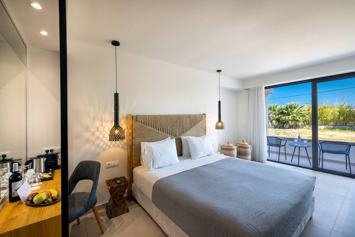 Una habitación de hotel moderna con una cama grande, un área de comedor con frutero y un balcón con mobiliario exterior.