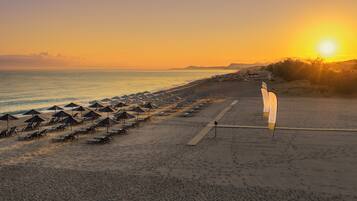 Uma praia com fileiras de espreguiçadeiras e guarda-sóis, um deck de madeira e uma vista clara do pôr do sol sobre o oceano.
