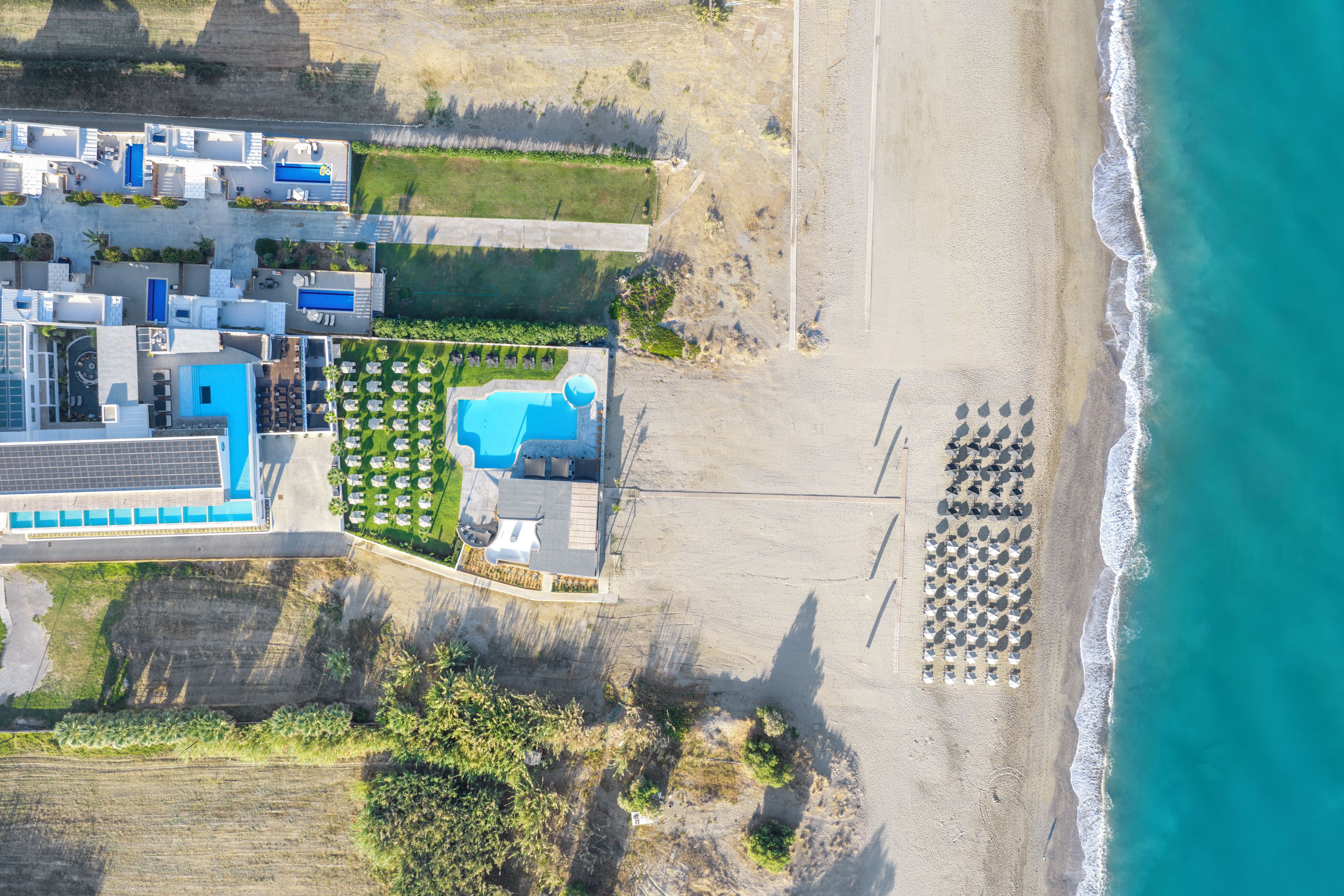 Vista aérea de un hotel costero con múltiples piscinas, una playa con sombrillas y un estacionamiento.