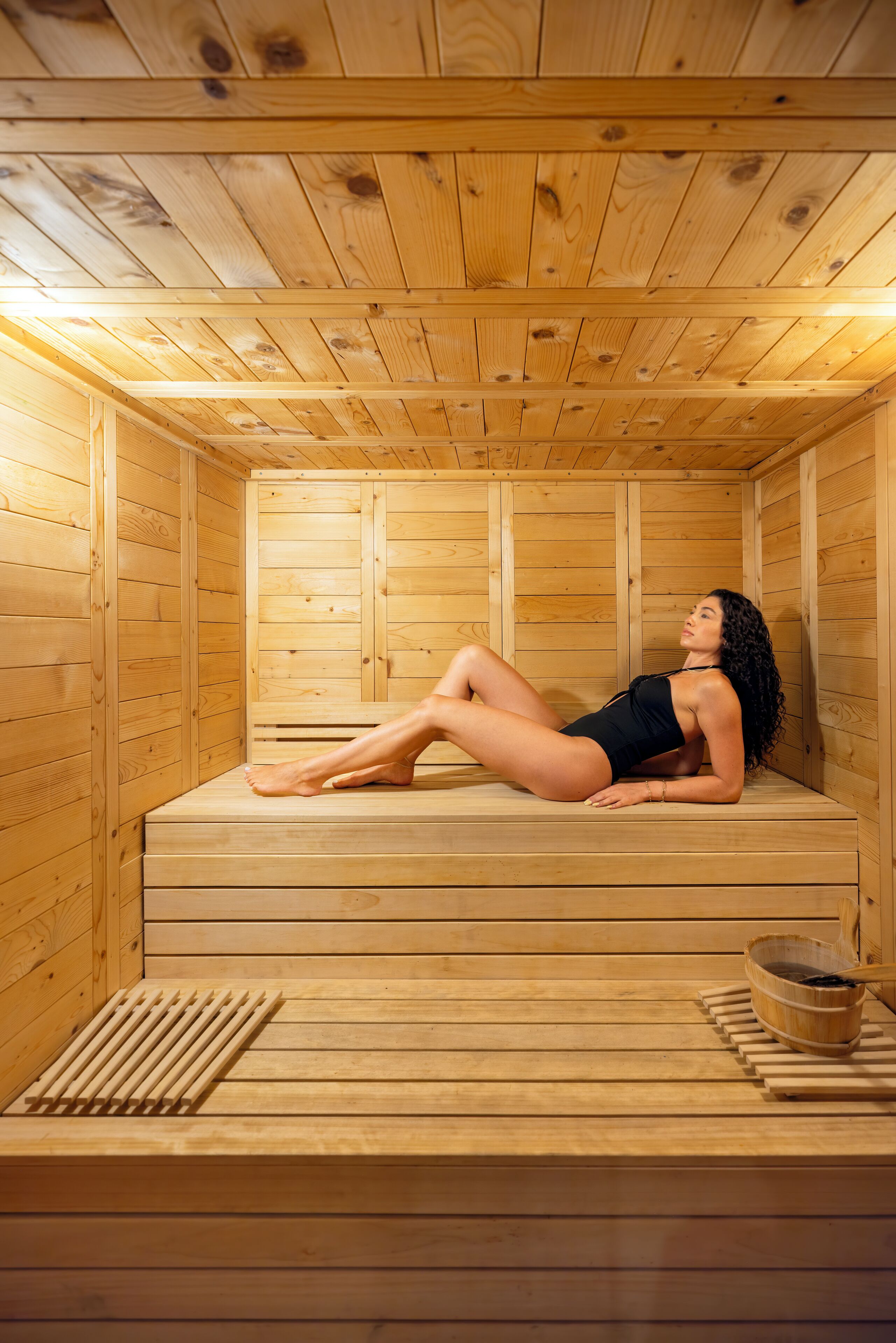 A sauna with wooden walls and benches, a person lying on a bench, and a bucket with a ladle.