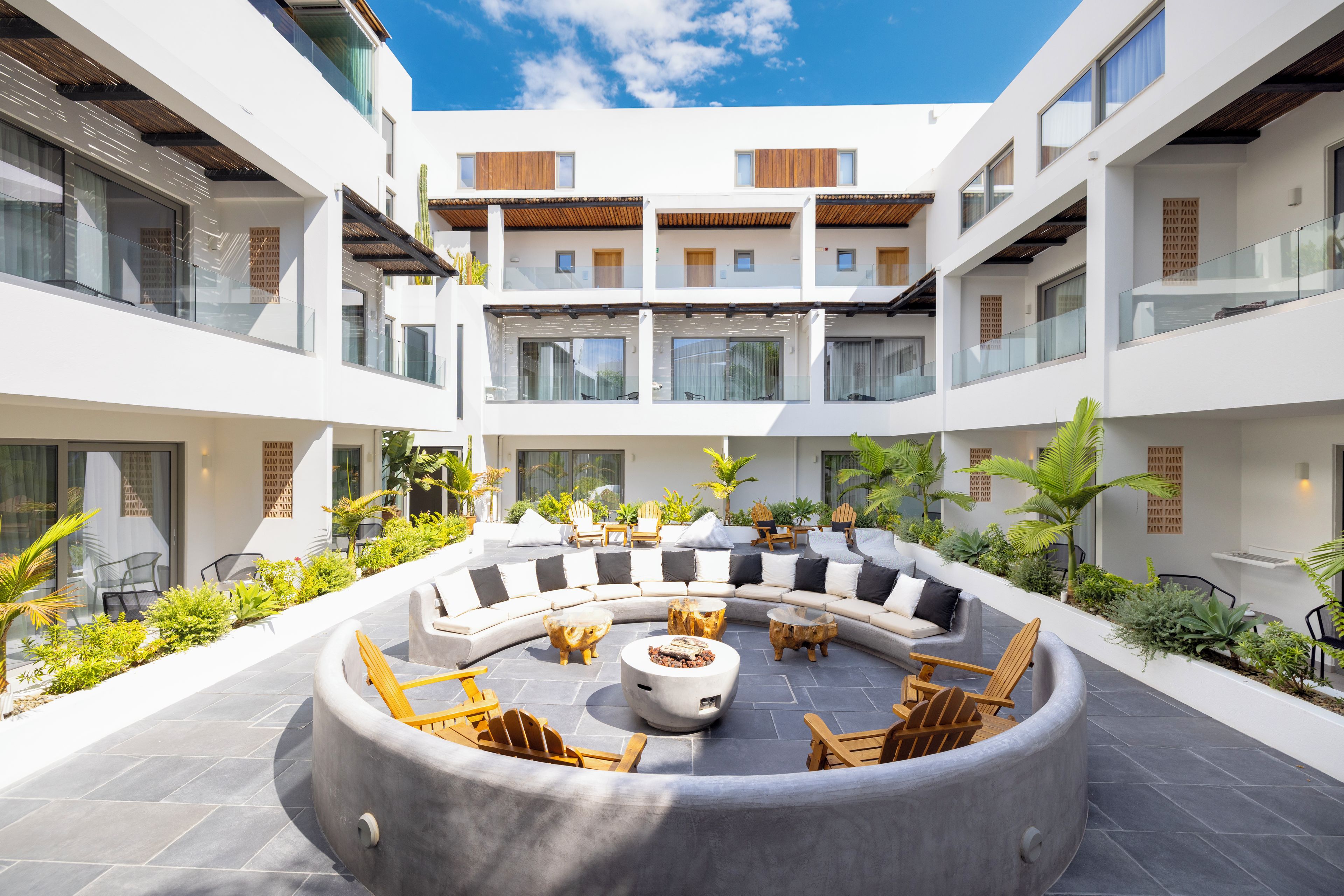 A modern outdoor seating area with a curved sofa and chairs, surrounded by potted plants and a fire pit.