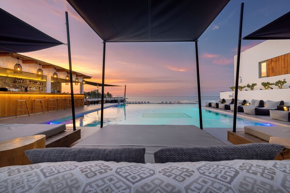 Un espace aménagé au bord de la piscine, avec un bar, des chaises longues et une vue sur la mer au coucher du soleil.