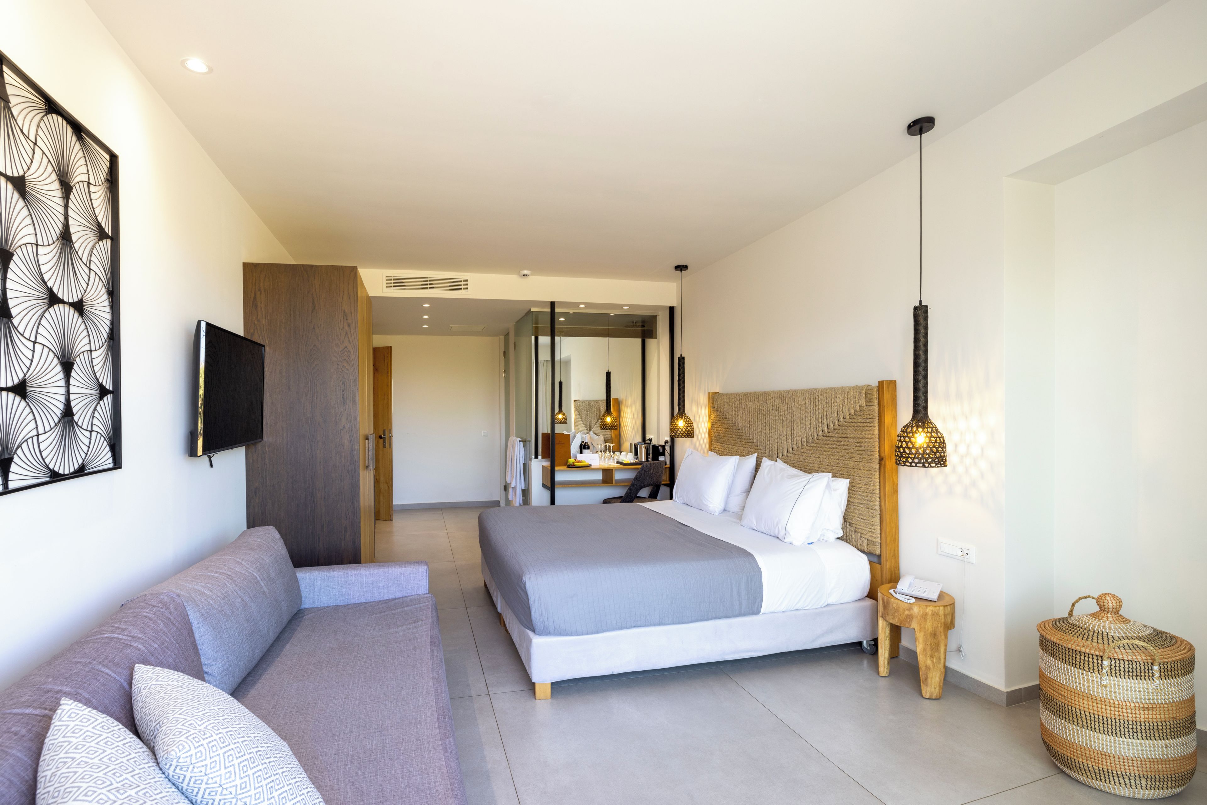 A modern hotel room with a large bed, a sofa, a TV, and a dining area in the background.