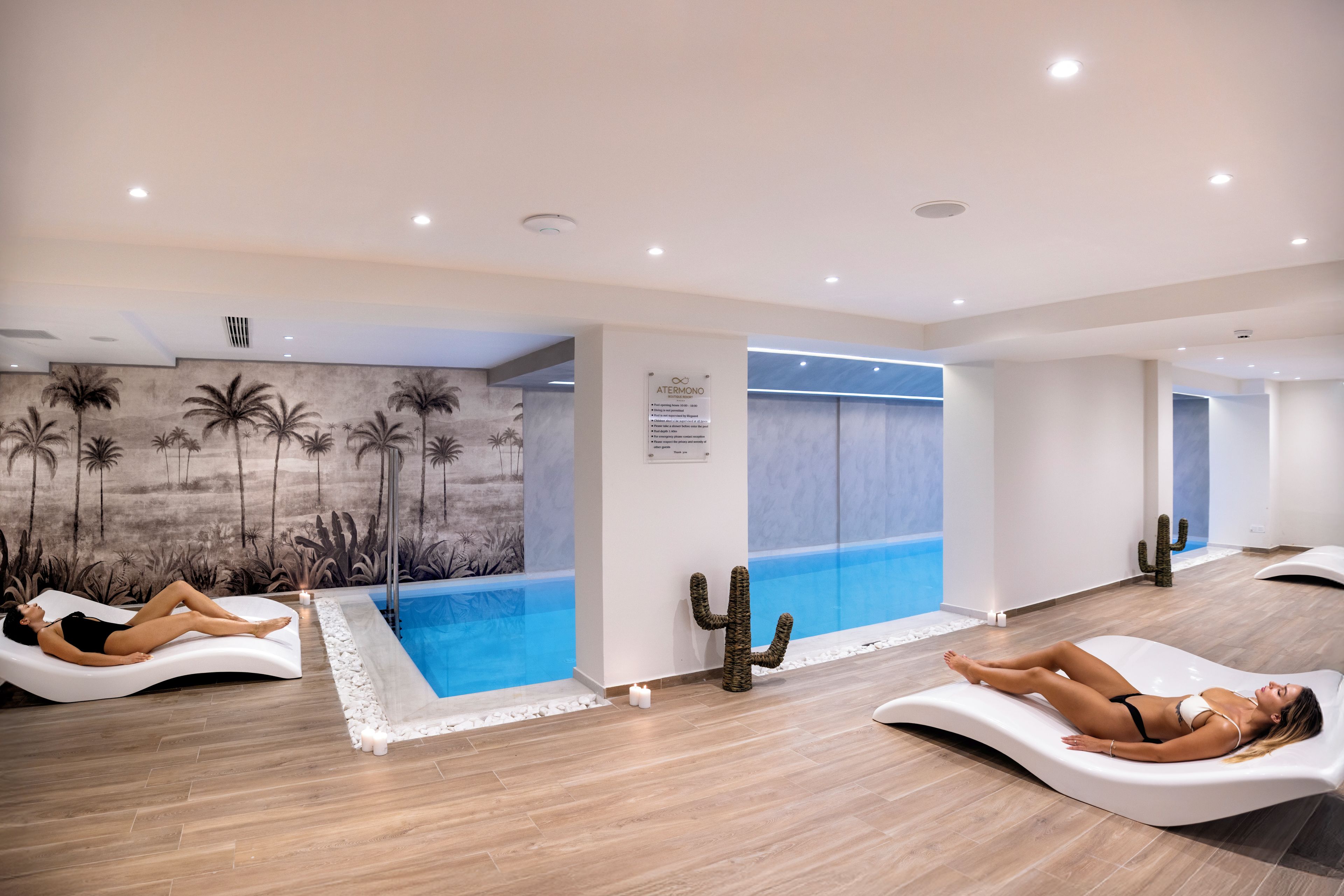 A modern spa area with two reclining massage chairs, a wall mural of palm trees, and a small indoor pool.