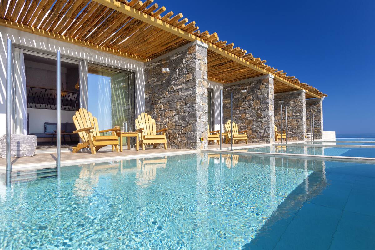 Zona junto a la piscina con sillas de madera y un muro de piedra, con cielo azul despejado de fondo.