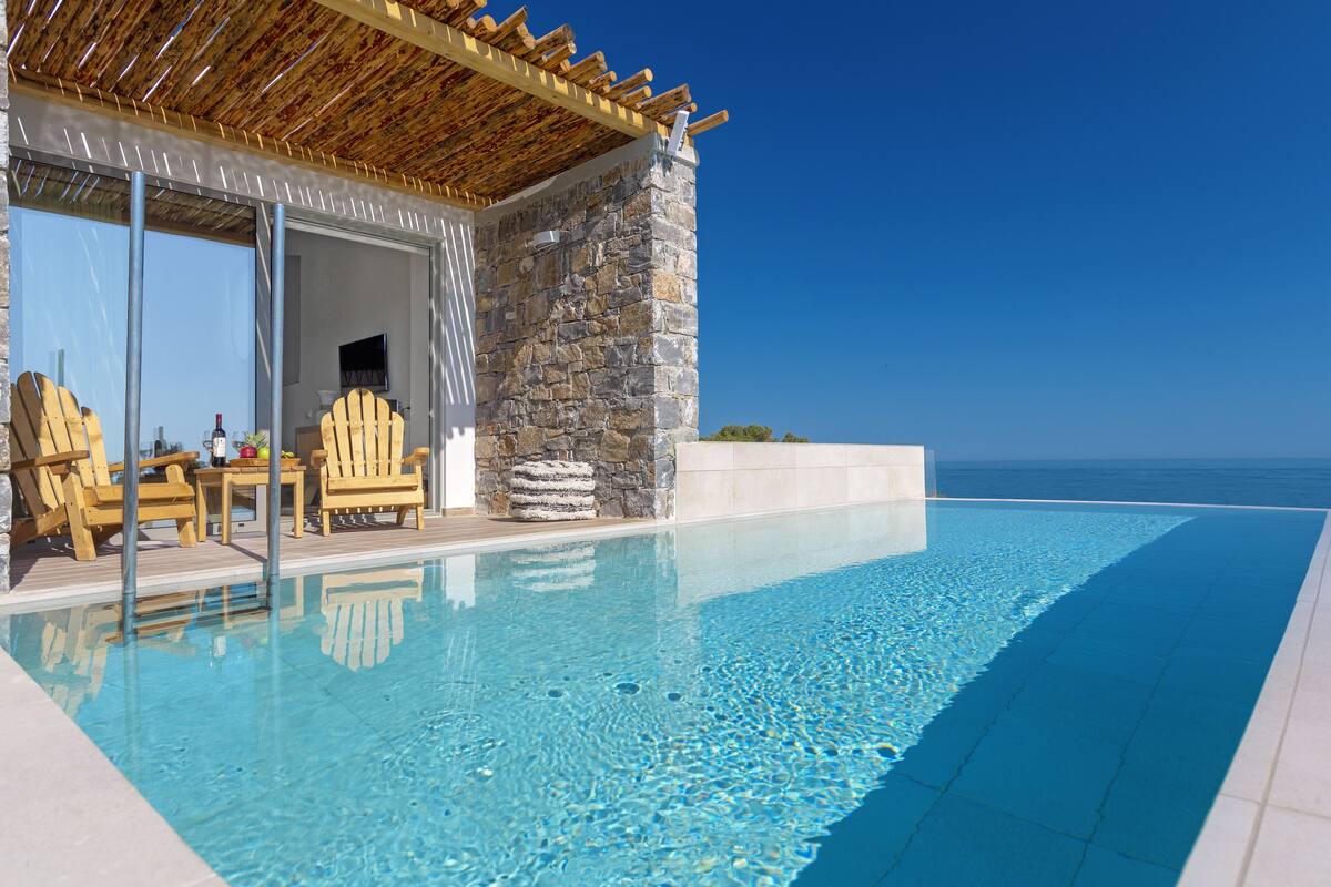 Zona junto a la piscina con pared de piedra, sillas de madera y una pérgola.