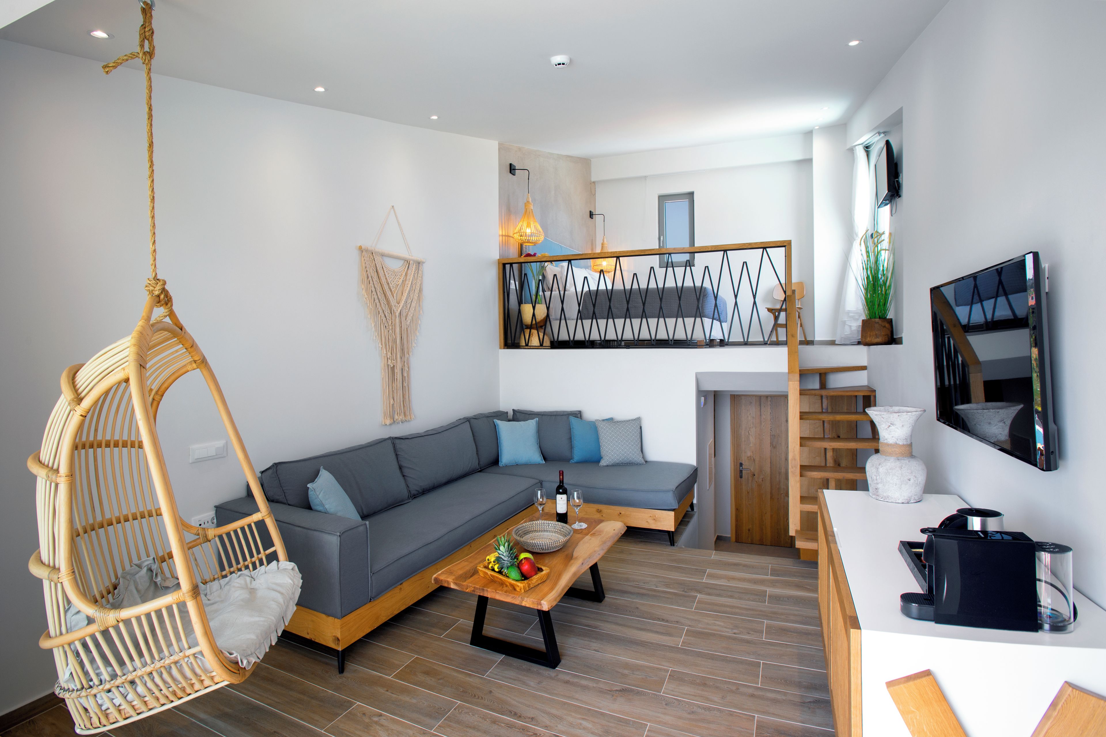 Ein modernes Wohnzimmer mit einem grauen Sofa, einem Hängekorbsessel, einem Holztisch und einem Fernseher an der Wand.