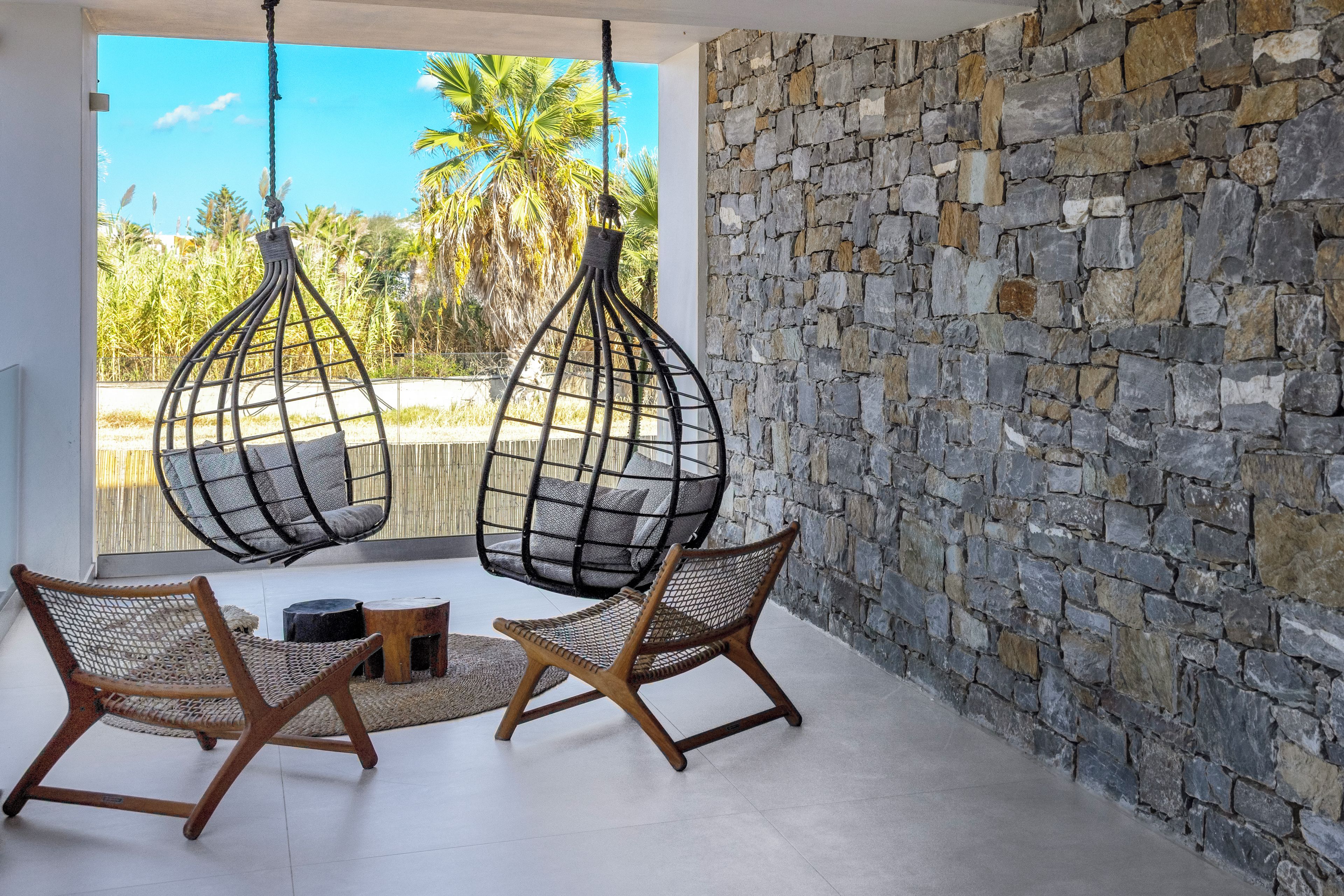 Una habitación con pared de piedra, dos sillas colgantes, una mesita y vistas a palmeras.