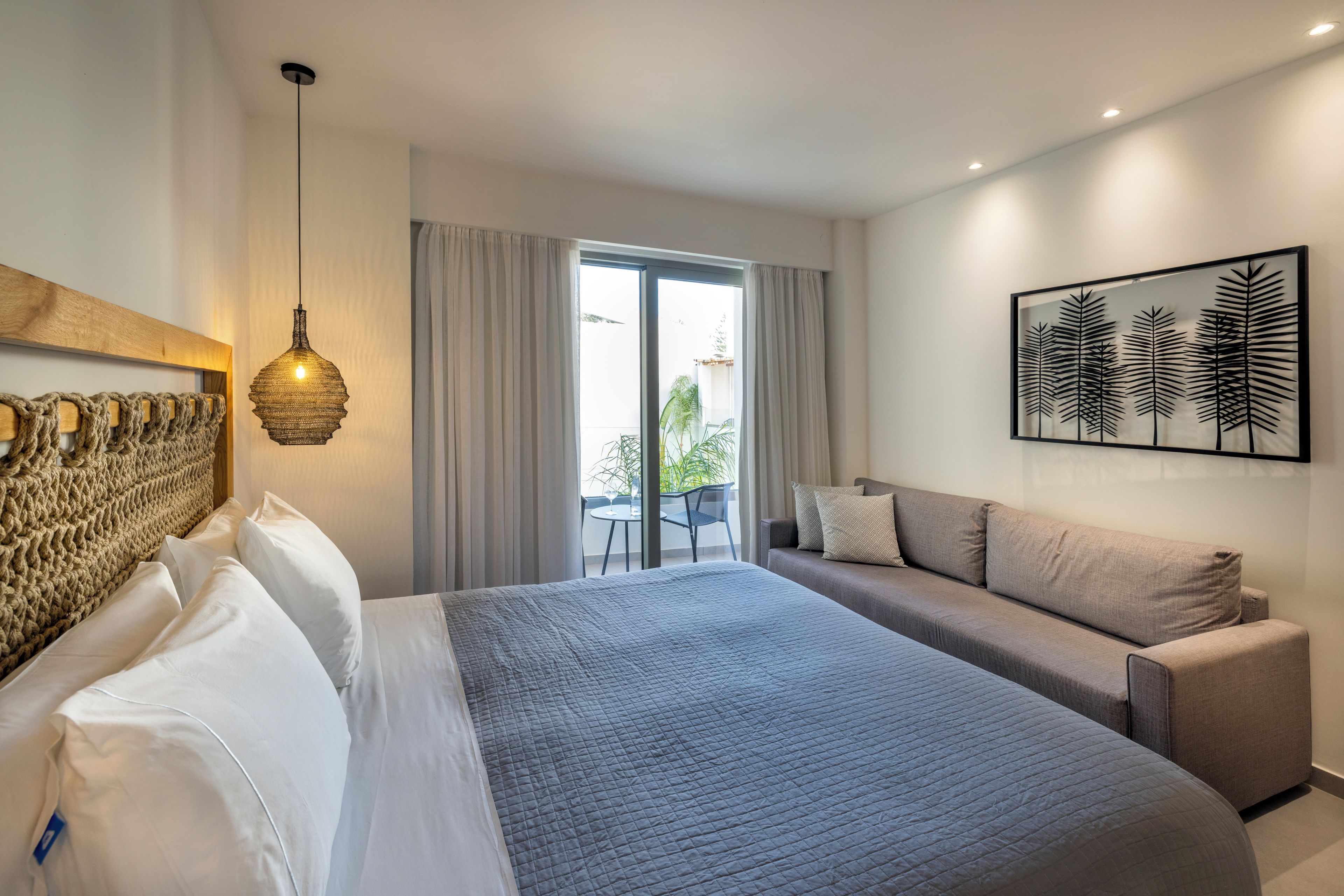 A modern hotel room with a large bed, a sofa, a dining area, and a view of the outdoors.