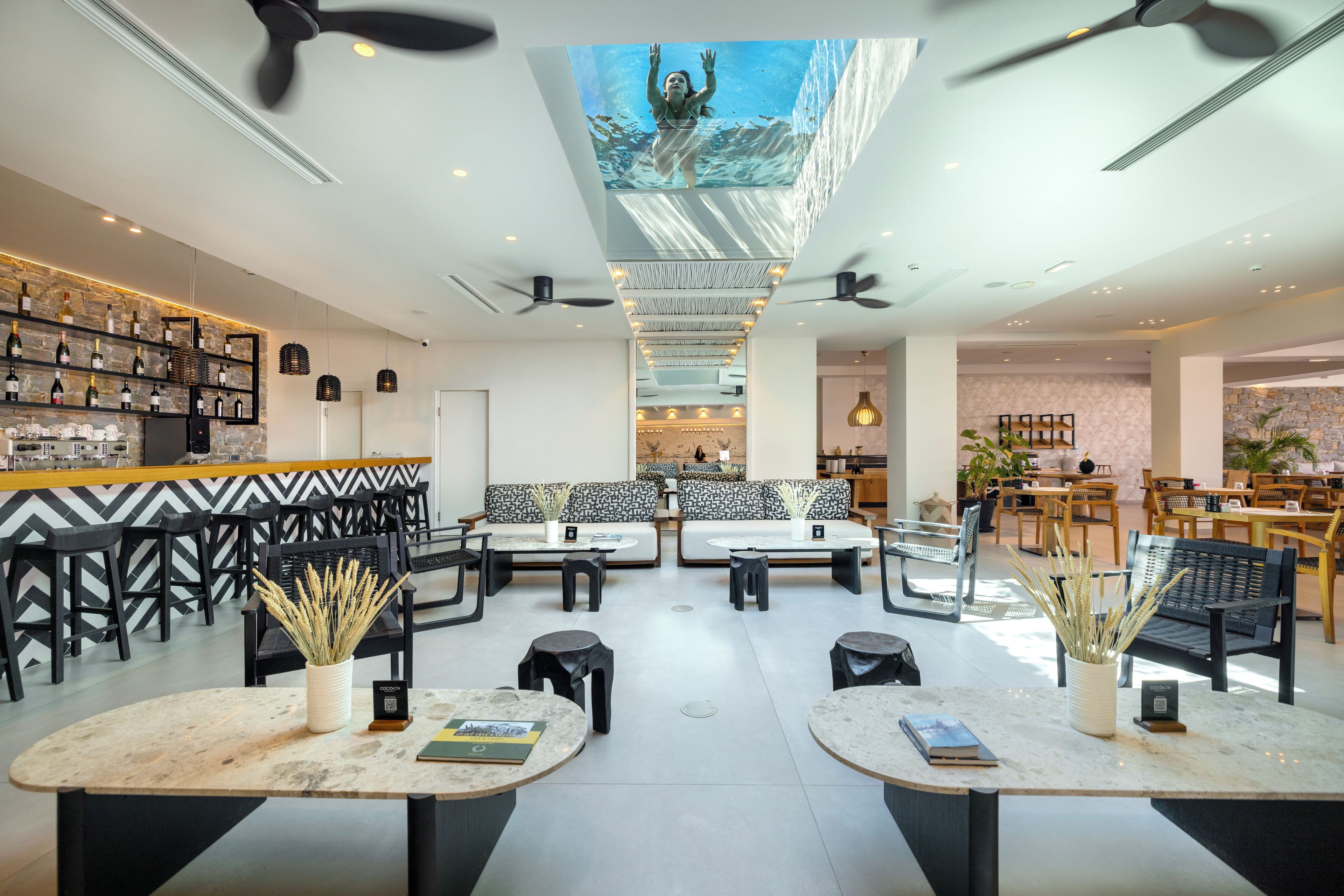 A modern interior with a bar area, seating with black and white patterned cushions, and a ceiling with a large window overlooking a pool.