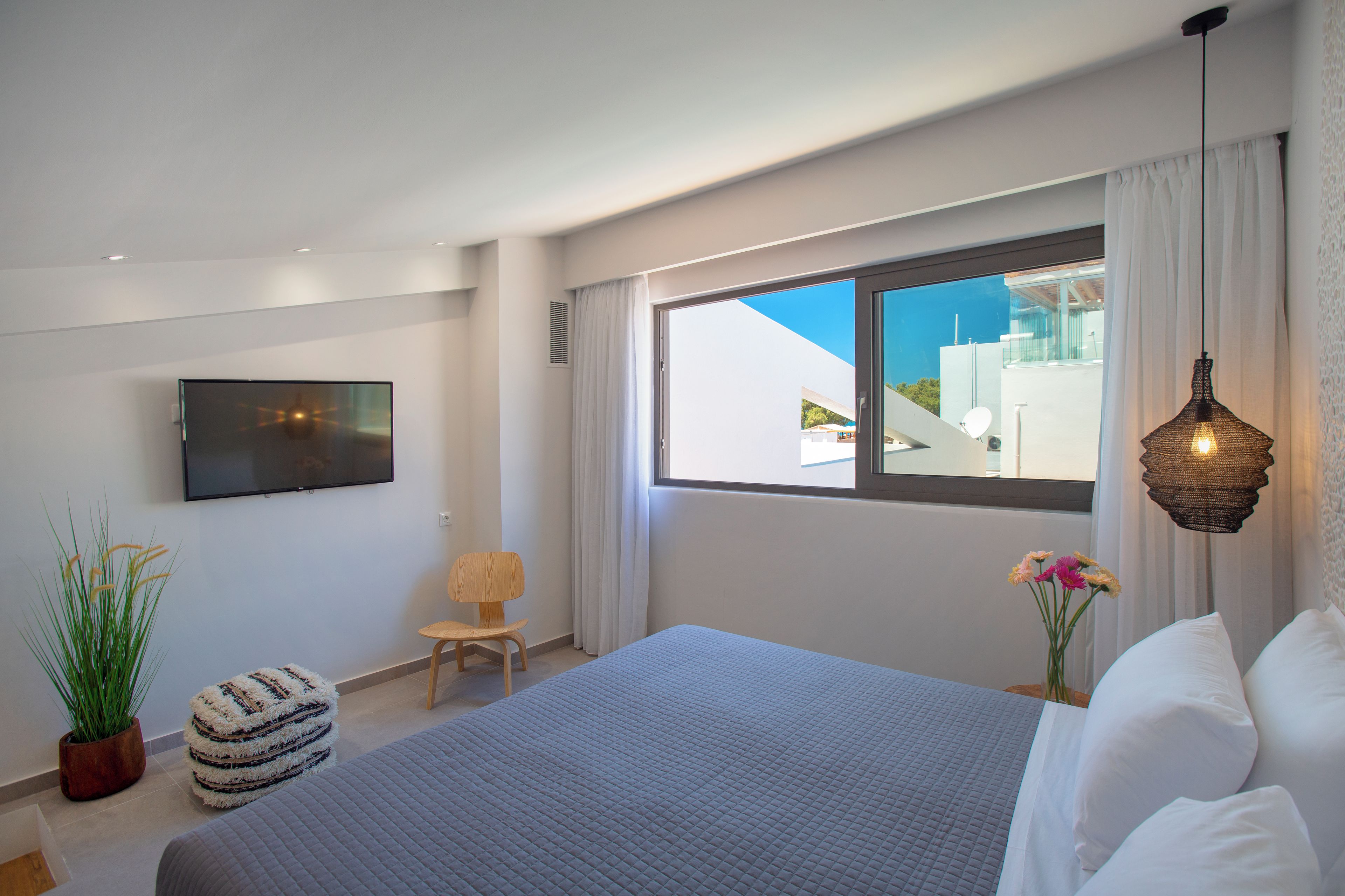 A modern bedroom with a large bed, a flat-screen TV, a potted plant, and a view of the outside.