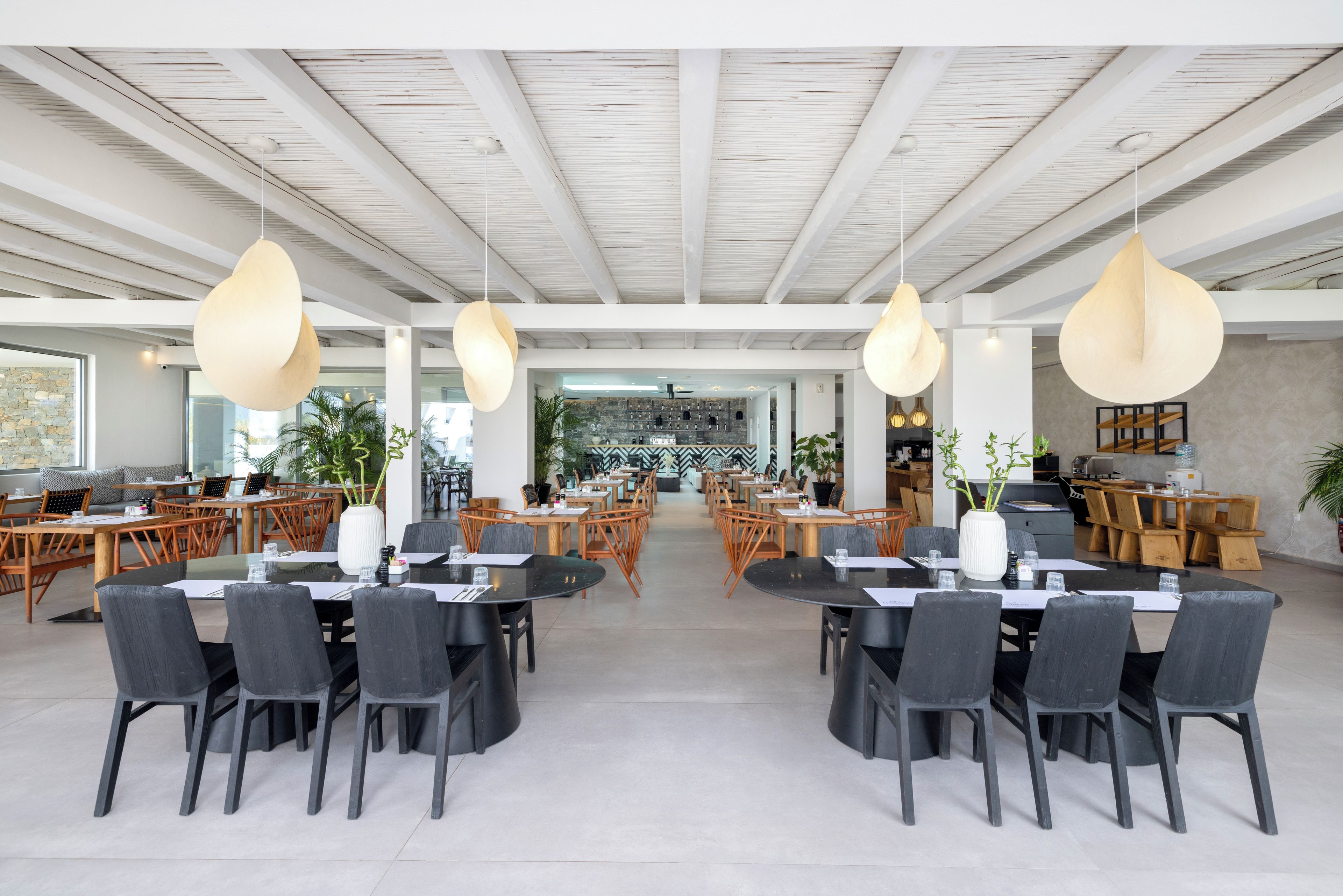 A spacious dining area with multiple tables and chairs, a kitchen in the background, and pendant lighting.