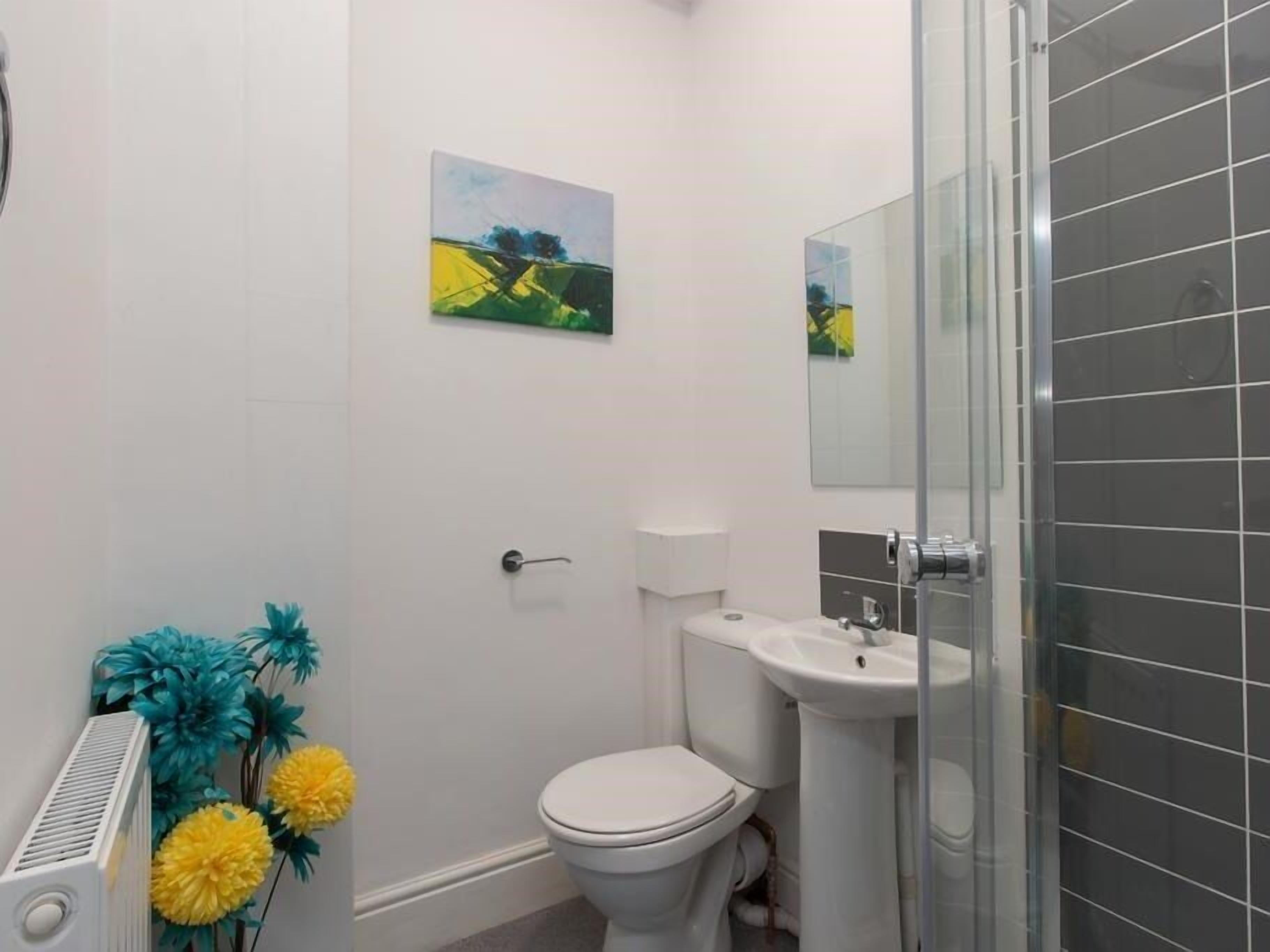 Baño con inodoro, lavabo y ducha. Hay dos cuadros colgados en la pared y un radiador decorado con flores artificiales.