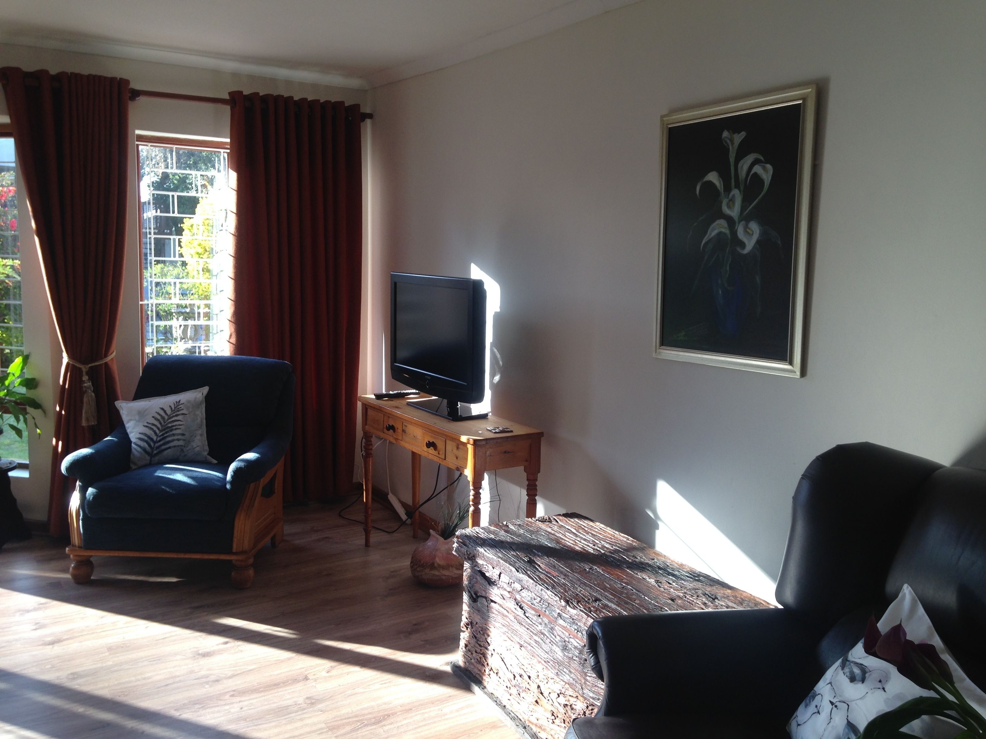 A living room with a television on a wooden stand, a blue armchair, a black leather sofa, and a painting on the wall.