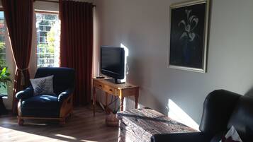 A living room with a television on a wooden stand, a blue armchair, a black leather sofa, and a painting on the wall.