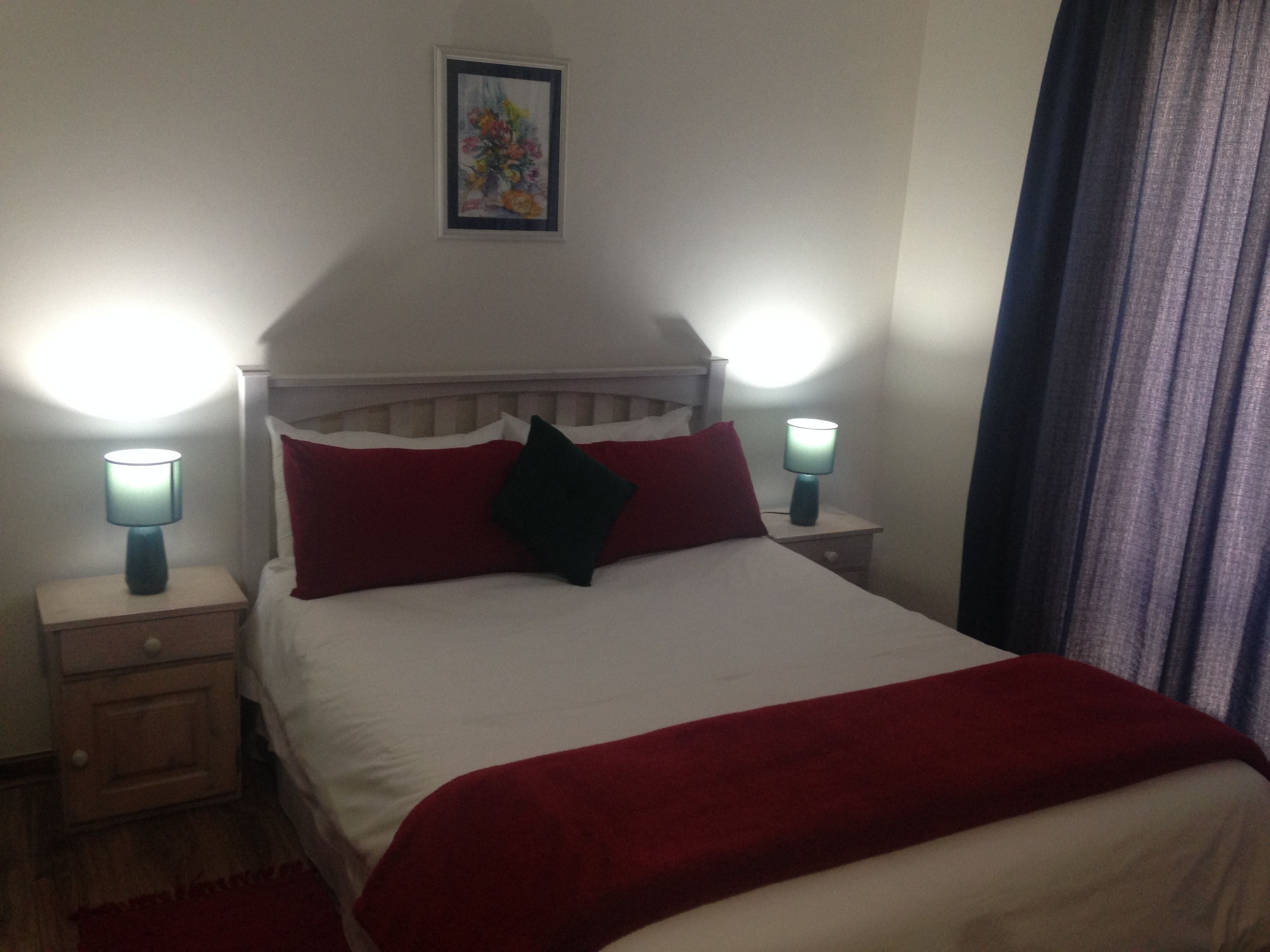 A neatly made bed with a white comforter and red pillows, flanked by two bedside tables with lamps, one of which is turned on.