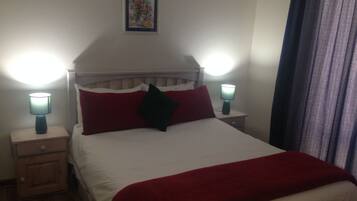 A neatly made bed with a white comforter and red pillows, flanked by two bedside tables with lamps, one of which is turned on.