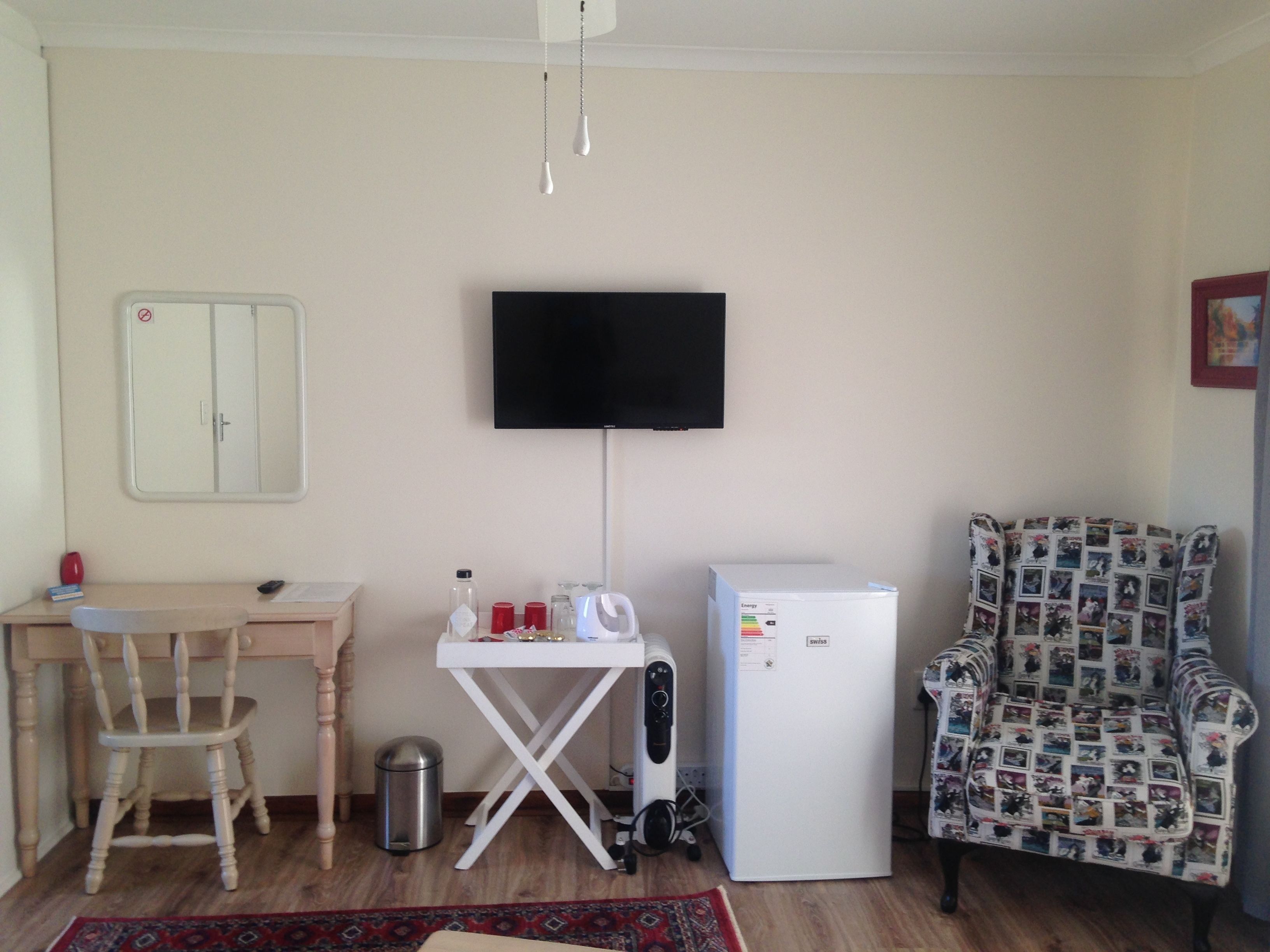 A small room with a desk, chair, TV, refrigerator, and patterned armchair.