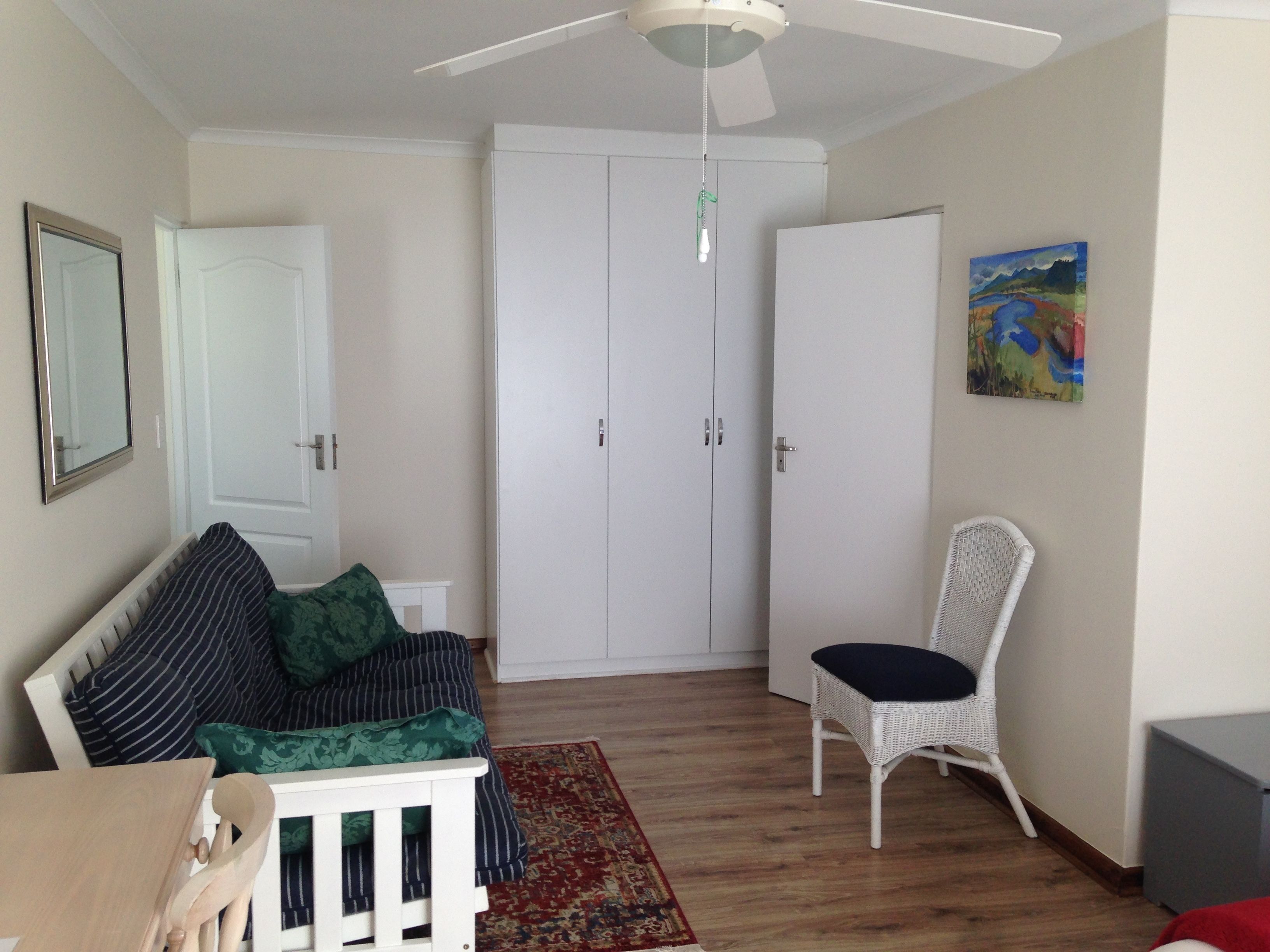 A room with a bed, a chair, a wardrobe, a ceiling fan, and a painting on the wall.