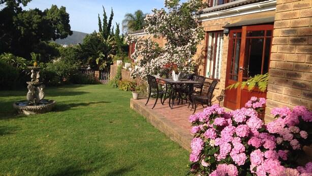 A well-maintained garden with a fountain, a patio table with chairs, and blooming flowers. - Blue Fern of Knysna (Knysna)