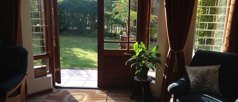 A room with a large sliding glass door leading to a garden, a wooden floor, a green sofa, and a small table with a plant.