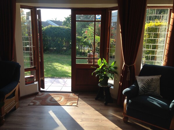A room with a large sliding glass door leading to a garden, a wooden floor, a green sofa, and a small table with a plant.