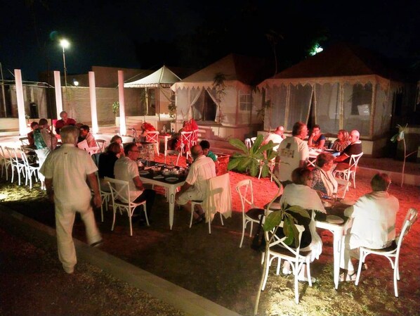 Un área de comedor al aire libre con mesas y sillas, rodeada por un muro bajo y un edificio con sección cubierta.
