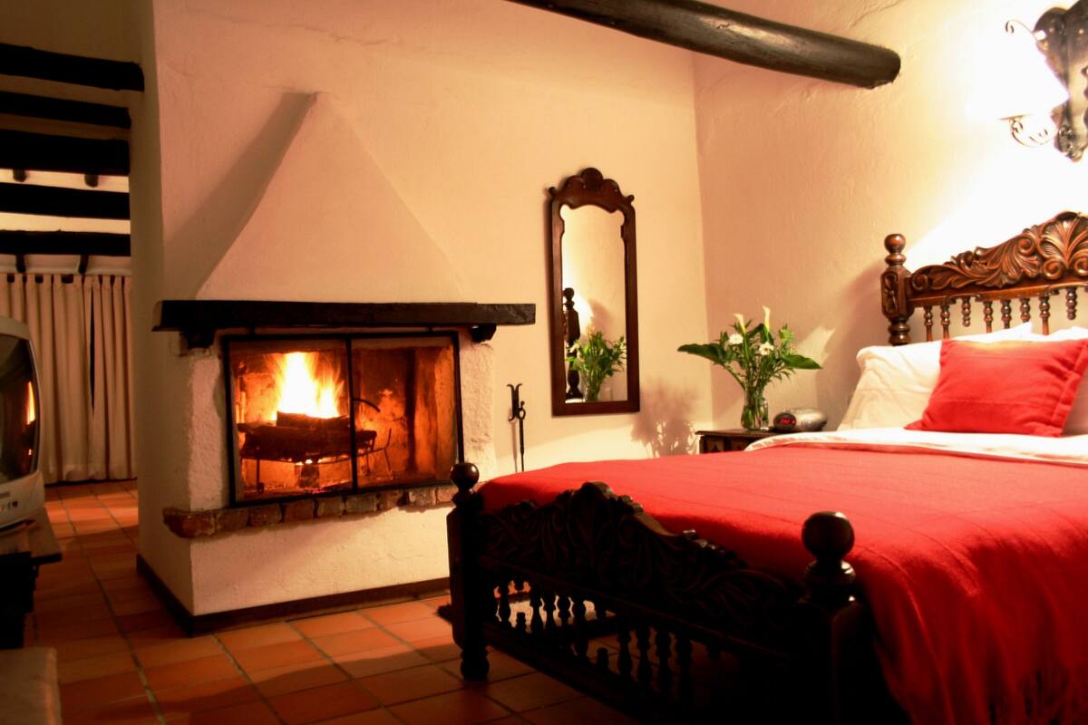 Un dormitorio con chimenea, una cama con colcha roja y un espejo en la pared.