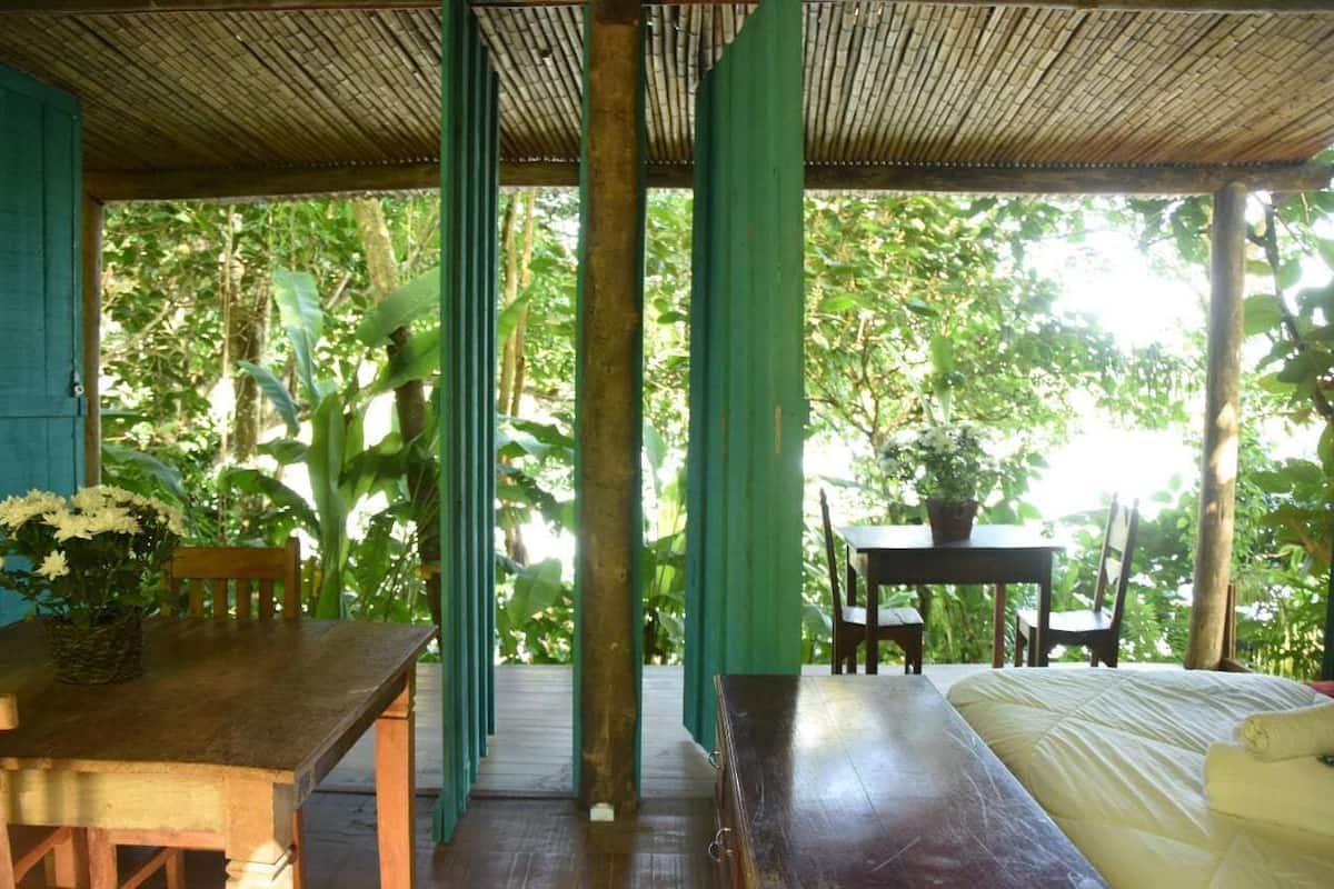 A wooden table with chairs and a bed in a covered outdoor area with a view of dense foliage.