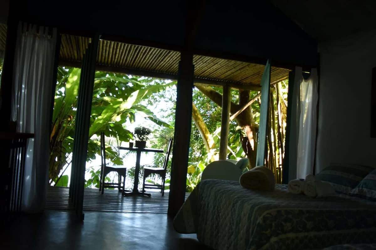 A room with a bed, a table with chairs, and a view of lush greenery outside.