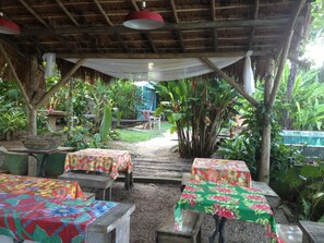 Un área de comedor al aire libre con mesas cubiertas con manteles florales, rodeada de abundante vegetación y una estructura de techo de paja.
