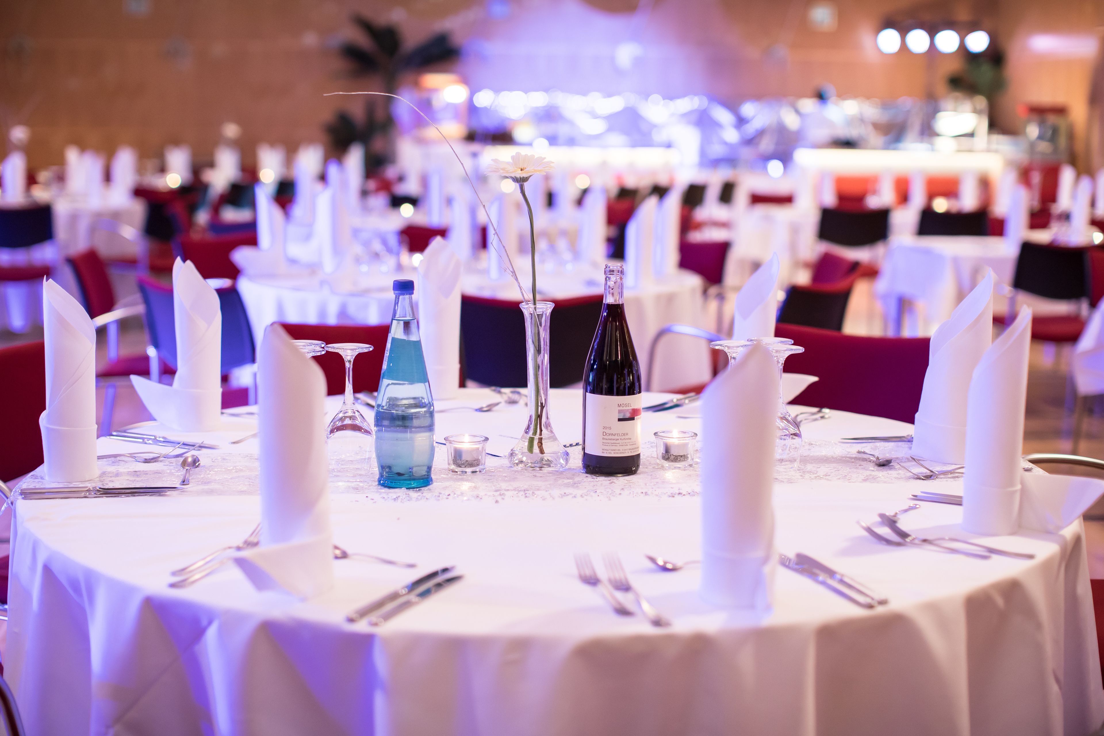 Een feestzaal met ronde tafels gedekt voor een diner, voorzien van witte tafellakens, servetten en bestek.
