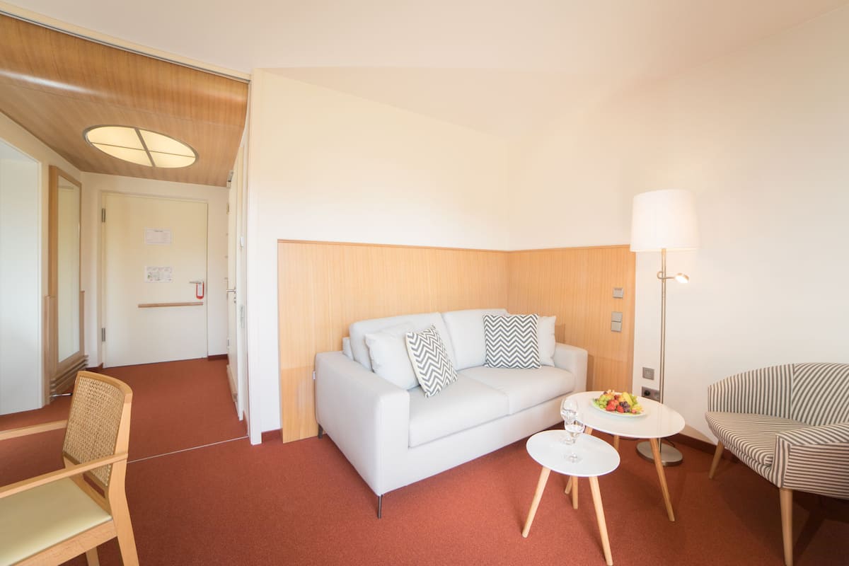 A modern hotel room with a white sofa, a small round table with a fruit bowl, a striped armchair, and a standing lamp.