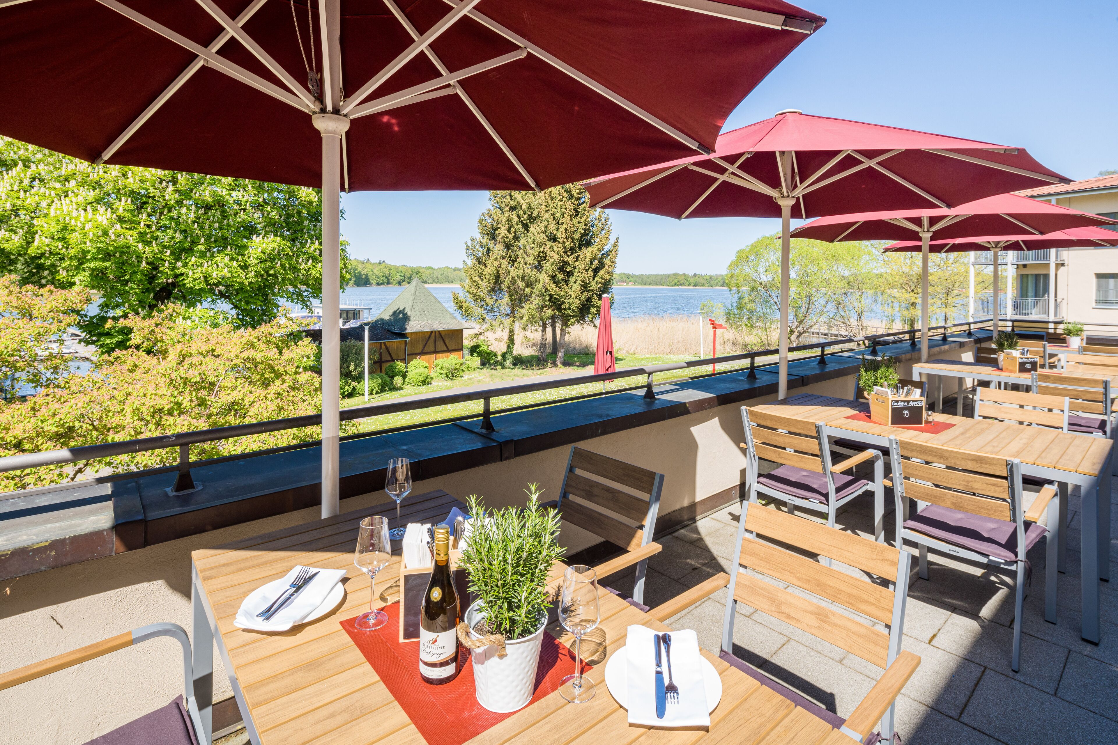 An outdoor dining area with wooden tables and chairs, red umbrellas, and a view of a lake and trees.