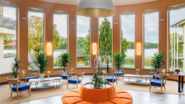 Eine geräumige Lobby mit großen Fenstern, einer zentral gelegenen orangefarbenen Sitzgruppe und zahlreichen Blumentöpfen.