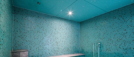 A bathroom with a tiled wall, a bench, a shower, and a drain.