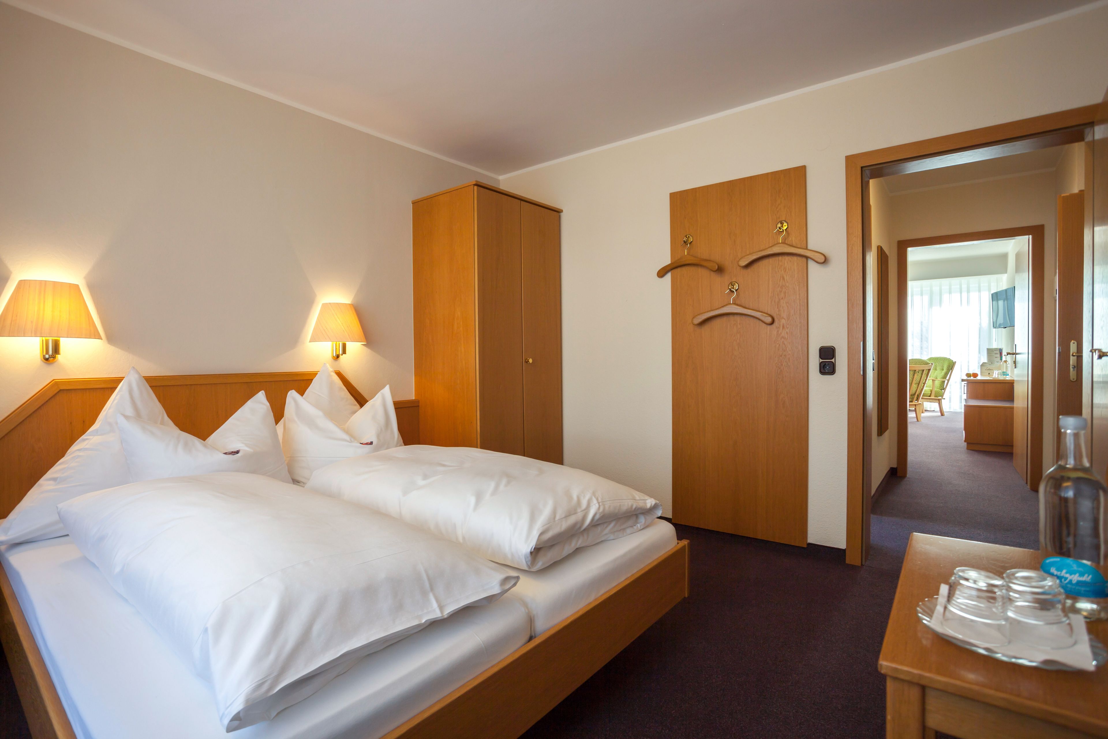 A hotel room with a double bed, two bedside lamps, a wooden wardrobe, a small table with a bottle and glasses, and a mirror.