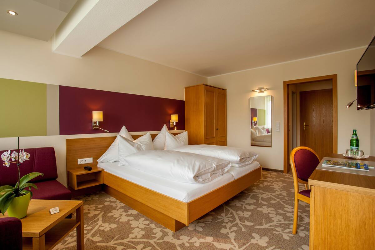 A hotel room with a large bed, a wooden desk, a chair, a small table, a potted plant, and a mirror on the wall.
