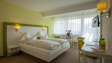 Une chambre d’hôtel comprenant un lit, des tables de chevet, un bureau avec une chaise, une télévision et une fenêtre avec des rideaux légers.