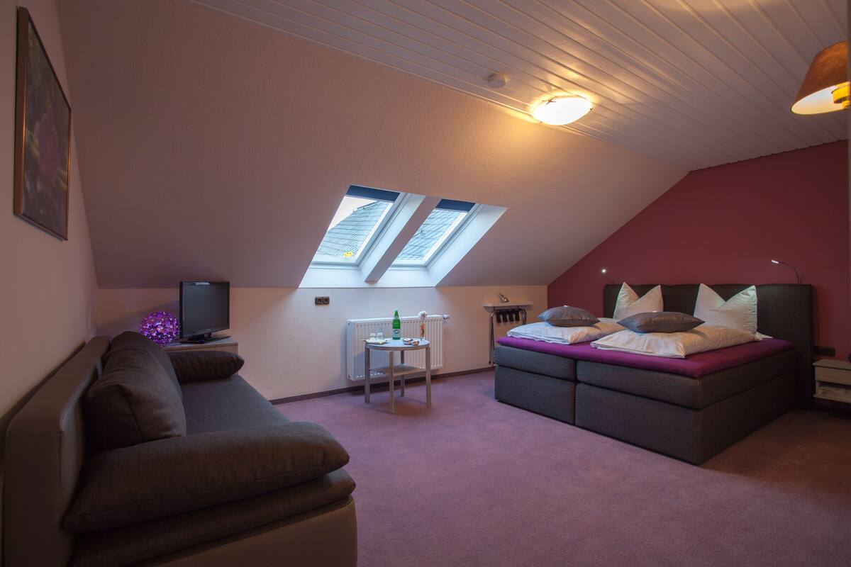 A bedroom with a sloped ceiling, a bed with multiple pillows, a small table, a radiator, and a television.