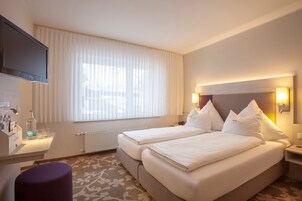 Una habitación de hotel con dos camas, televisión, escritorio con lámpara y ventana con cortinas.