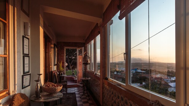A corridor with checkered flooring, wicker furniture, and large windows offering a view of a town and hills. - Shepinetree - Pinheira House (Sintra)