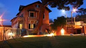 A two-story house with a visible front door and windows, surrounded by a well-maintained lawn and street lamps. - Shepinetree - Pinheira House (Sintra)