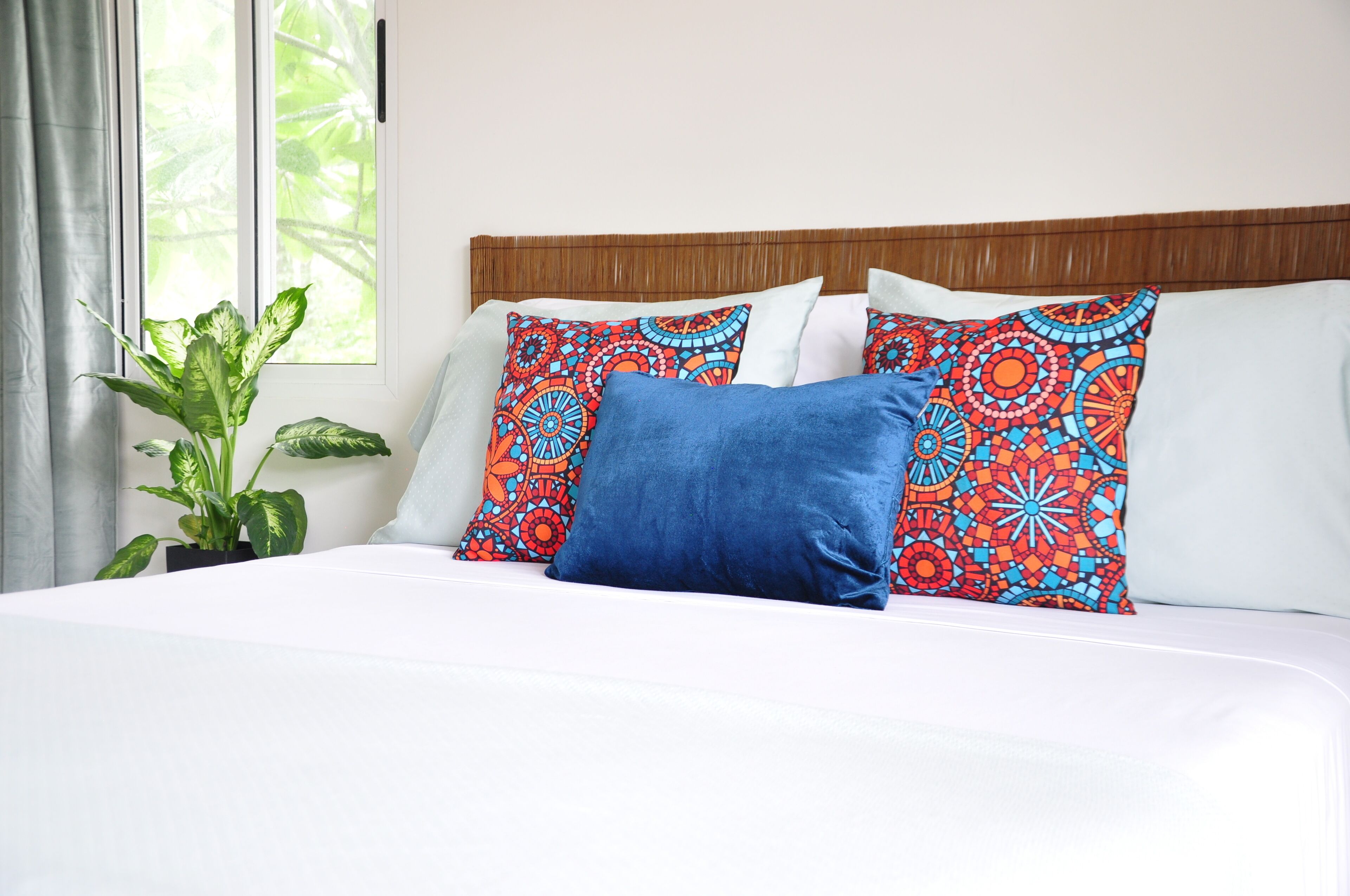 A bed with white bedding and colorful patterned pillows, a potted plant, and a window with curtains.