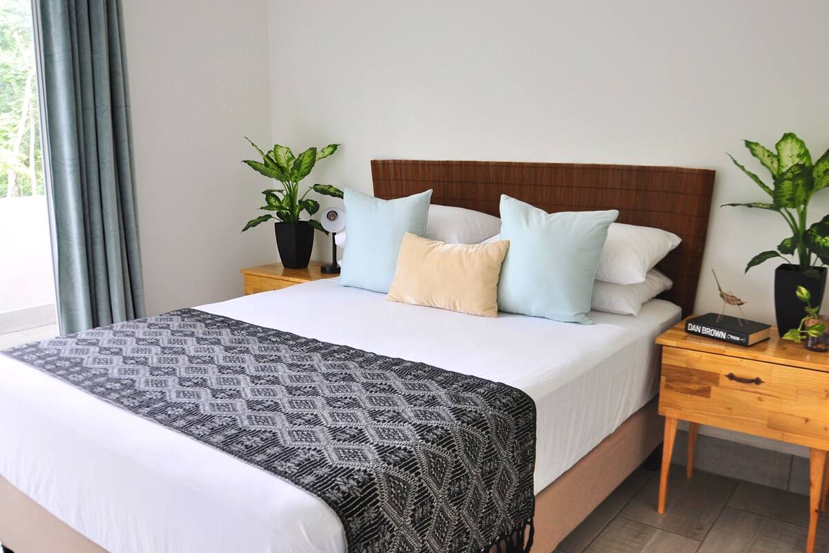 A neatly made bed with a patterned blanket, a wooden headboard, and multiple pillows. There are two bedside tables with potted plants and a book titled "Dan Brown".